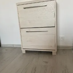 Dwupoziomowa szafka na buty z jasnego drewna z metalowymi uchwytami, ustawiona na podłodze imitującej drewno przy białej ścianie z gniazdkiem elektrycznym.