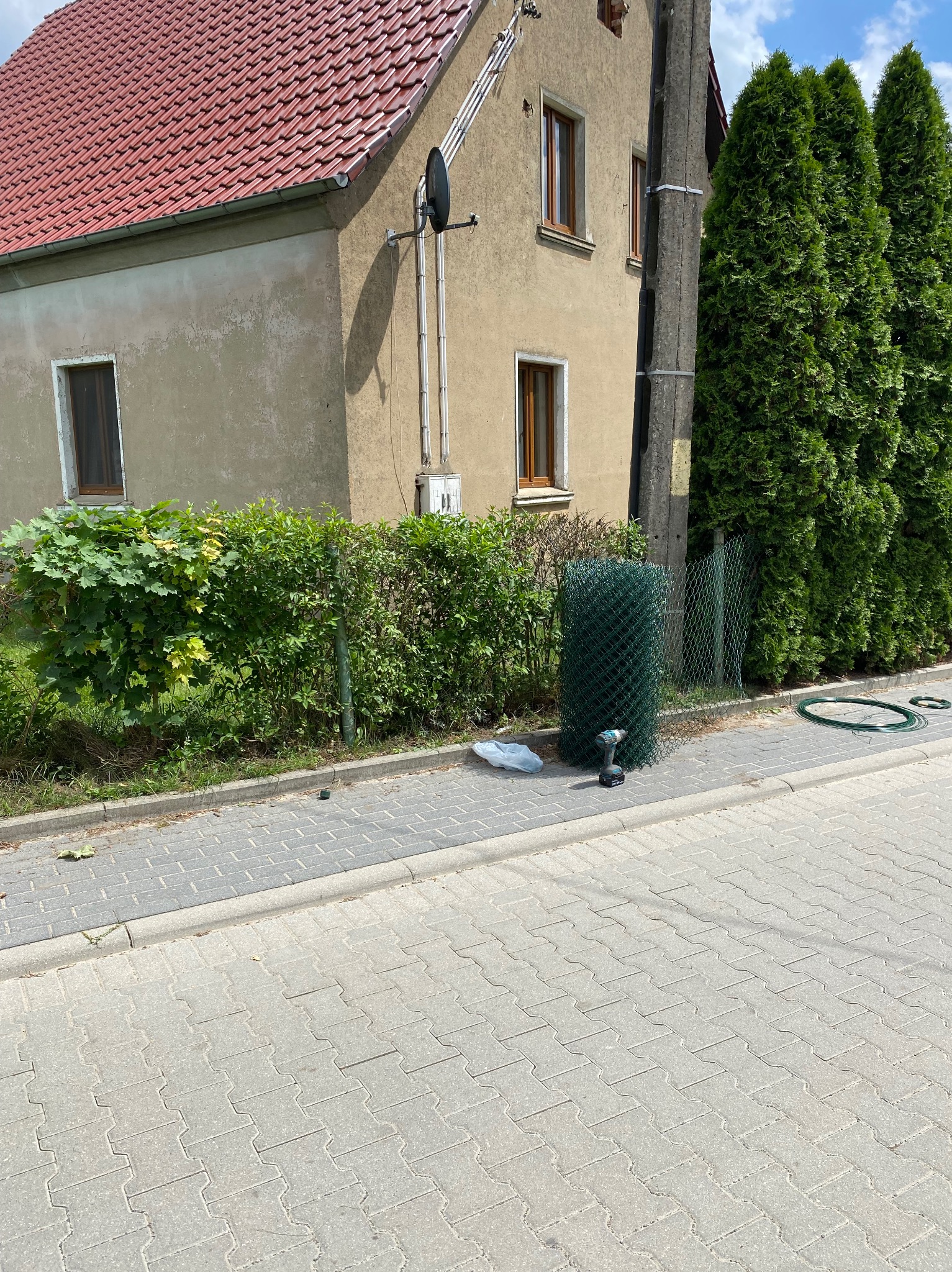 Zrolowana siatka ogrodzeniowa, wkrętarka akumulatorowa i zwój drutu leżą na chodniku obok domu, przygotowane do instalacji ogrodzenia.