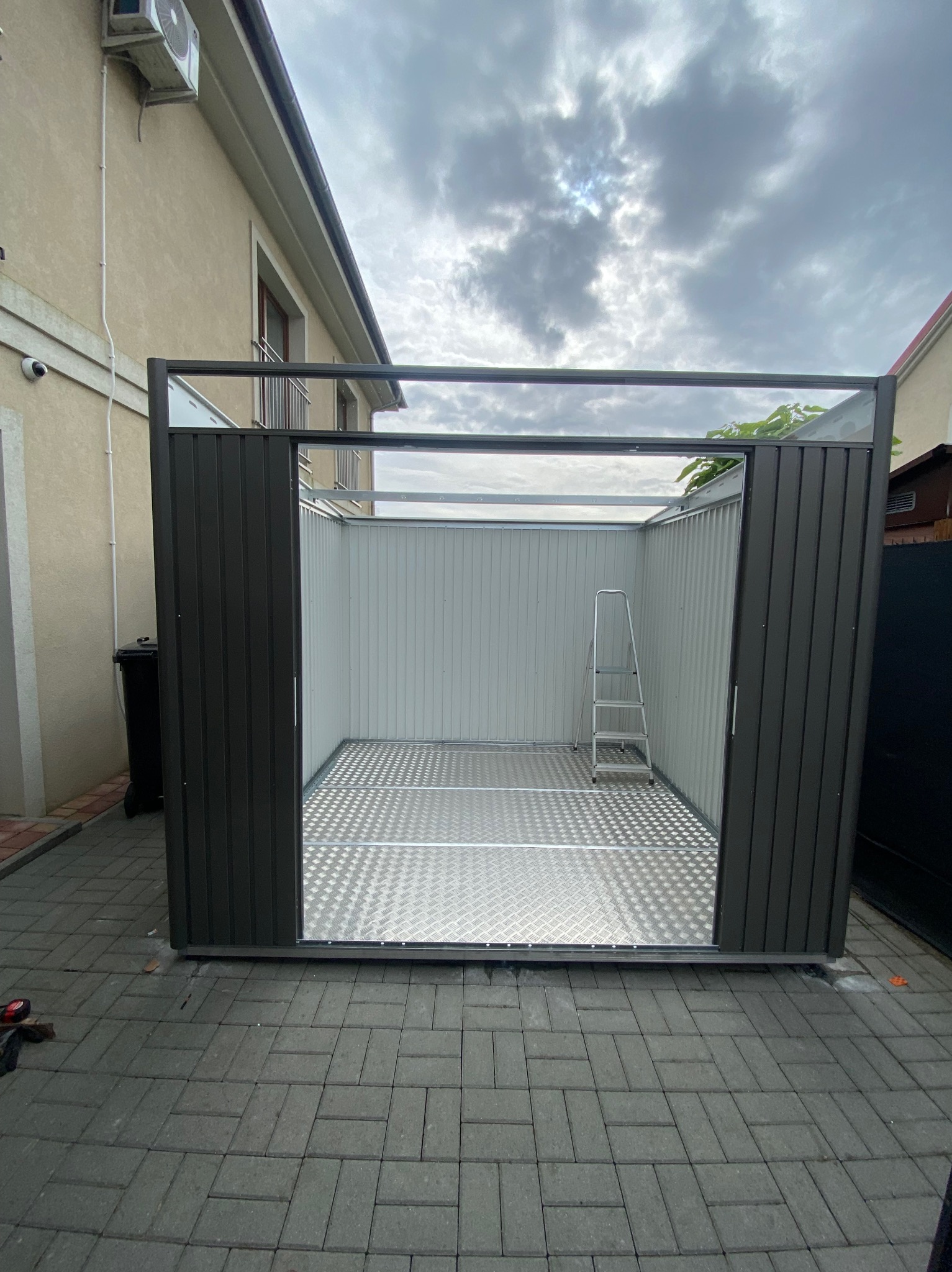 Nowoczesna, otwarta wiata z przesuwnymi drzwiami, metalową podłogą i drabinką wewnątrz, na tle brukowanej nawierzchni i budynku mieszkalnego.