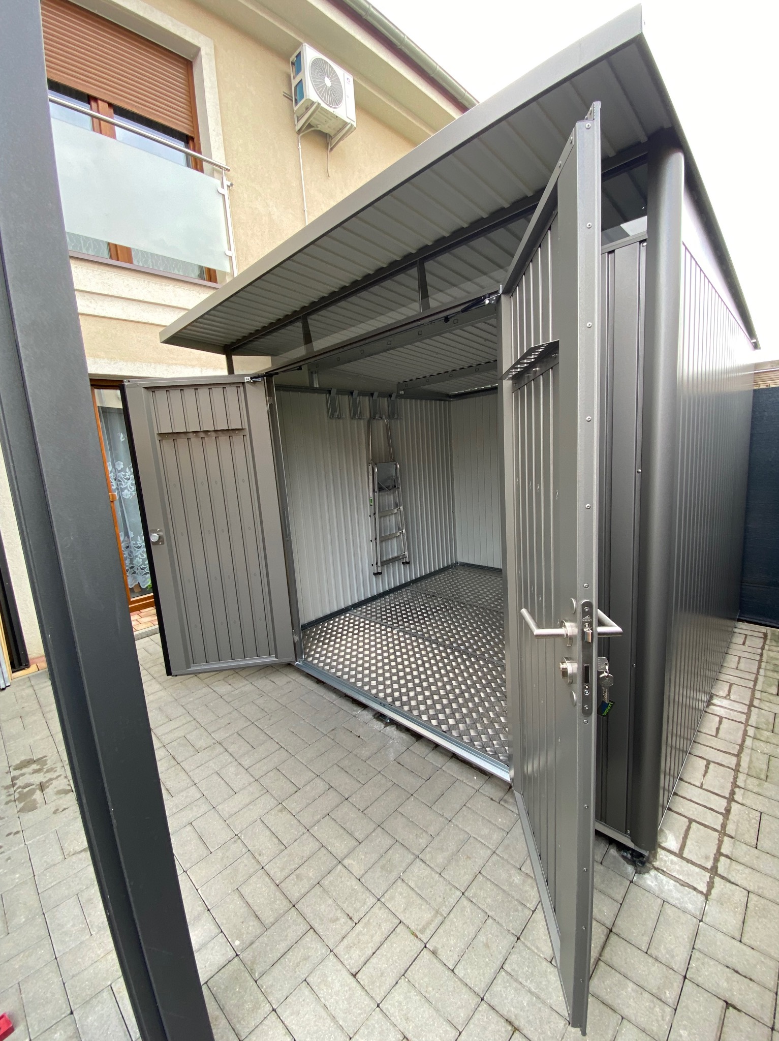 Szary metalowy schowek ogrodowy z otwartymi drzwiami, widoczna drabina wewnątrz, kostka brukowa na zewnątrz.