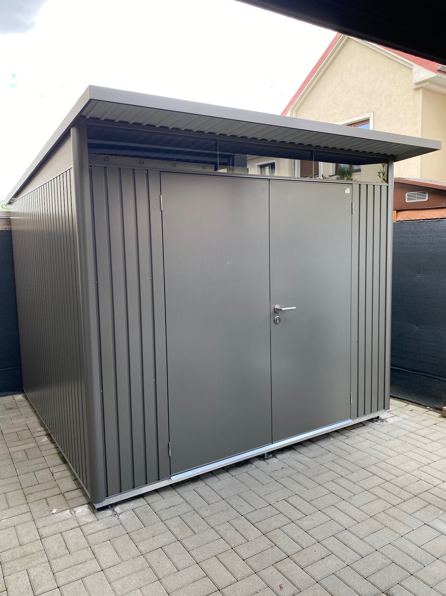 Nowoczesny, metalowy schowek ogrodowy z dwuskrzydłowymi drzwiami, umieszczony na kostce brukowej, z widocznym fragmentem domu w tle.