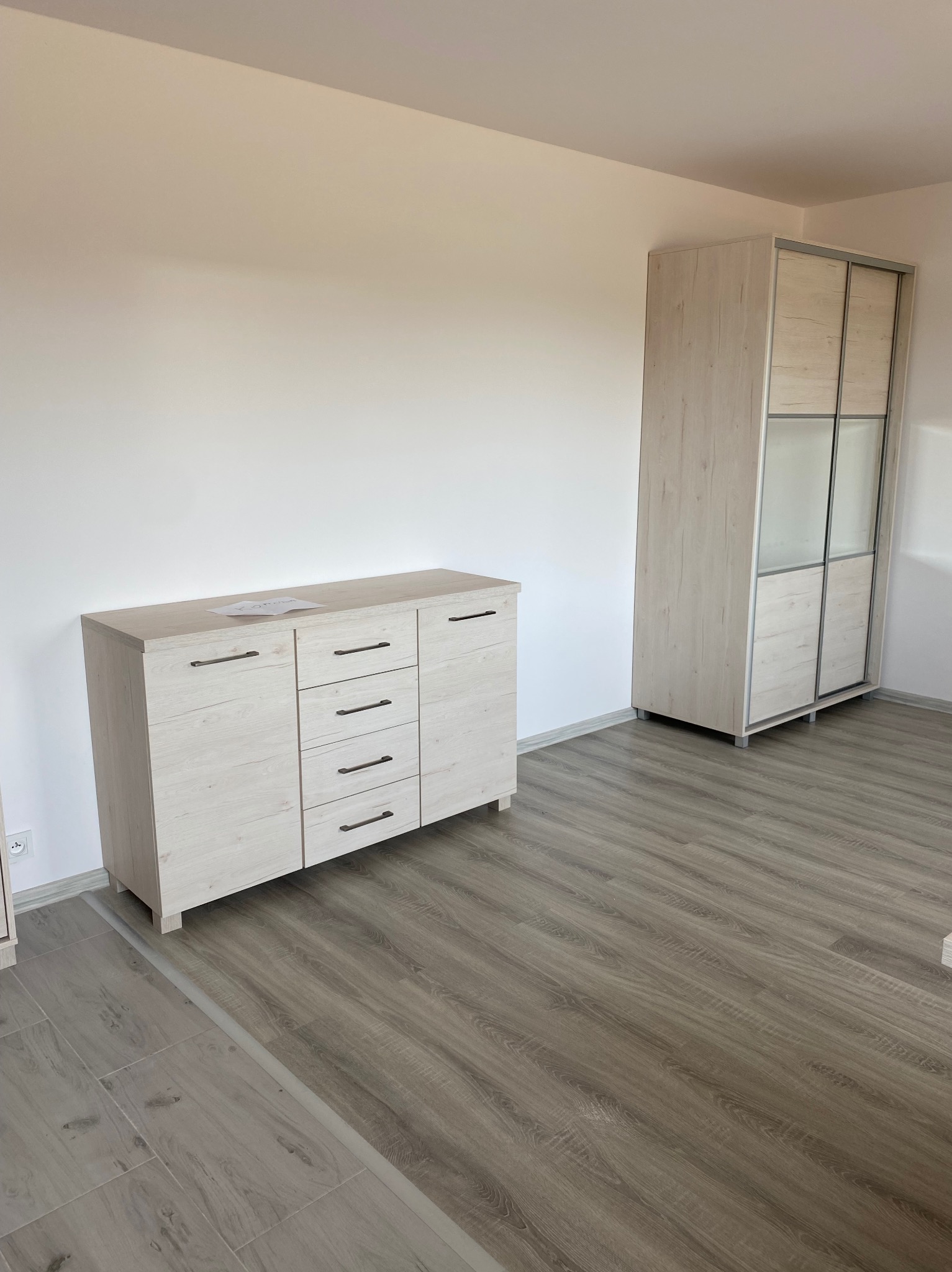 Jasna komoda z czterema drzwiczkami i pięcioma szufladami oraz przesuwna szafa wnękowa z lustrzanymi drzwiami, stojące na podłodze z paneli imitujących drewno w pustym pokoju.
