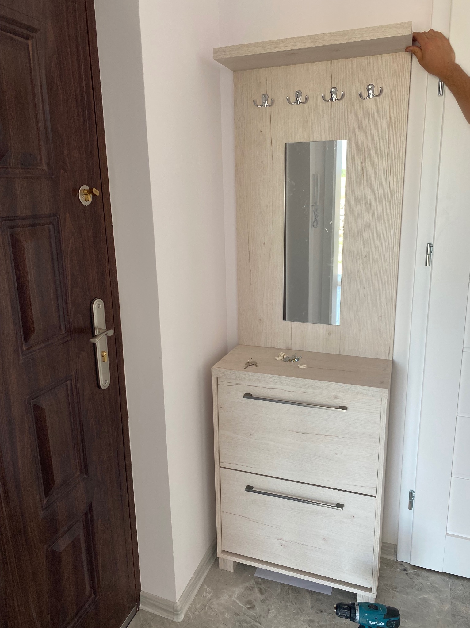 Nowoczesny przedpokój z lustrem i szafką na buty w jasnej kolorystyce, widoczny wiertarko-wkrętak akumulatorowy na podłodze, klucze i elementy montażowe na blacie szafki.