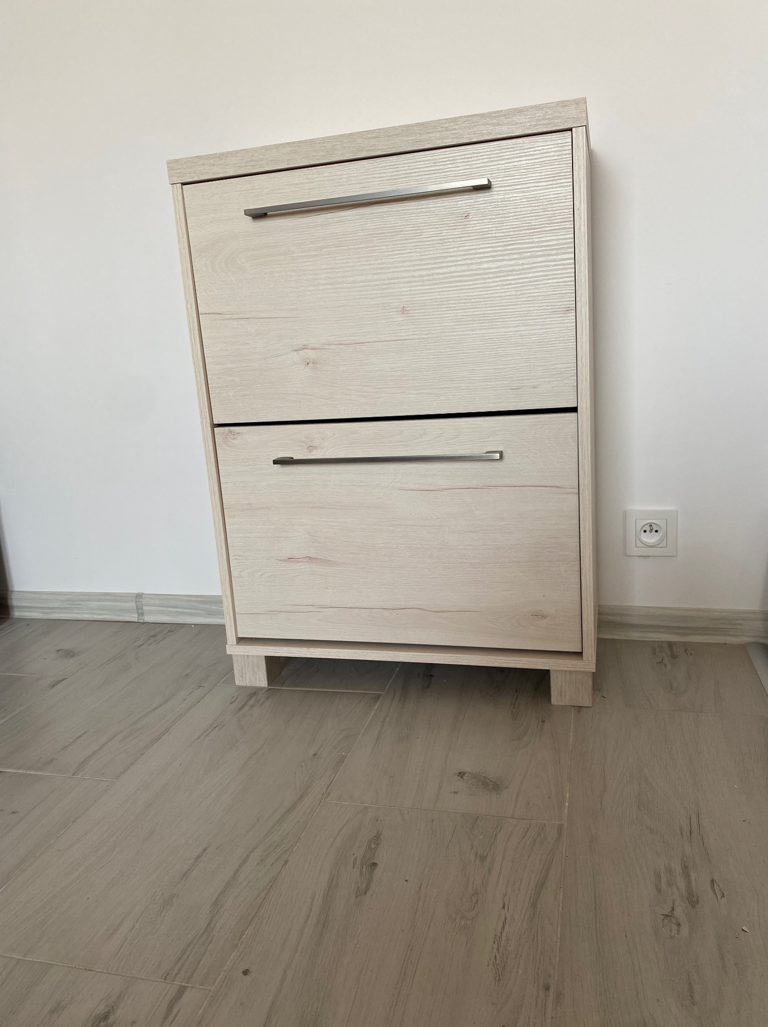 Dwupoziomowa szafka na buty z jasnego drewna z metalowymi uchwytami, ustawiona na podłodze imitującej drewno przy białej ścianie z gniazdkiem elektrycznym.
