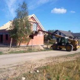 Budowa domu murowanego: Żółta ładowarka teleskopowa CAT podnosi bloczki na ścianę domu w trakcie budowy, widoczna więźba dachowa i częściowo wymurowane ściany, w tle inne budynki i błękitne niebo.