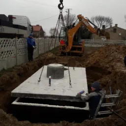 Montaż betonowego zbiornika pod ziemią przy użyciu koparki i dźwigu; pracownik na drabinie uszczelnia połączenie elementów zbiornika.