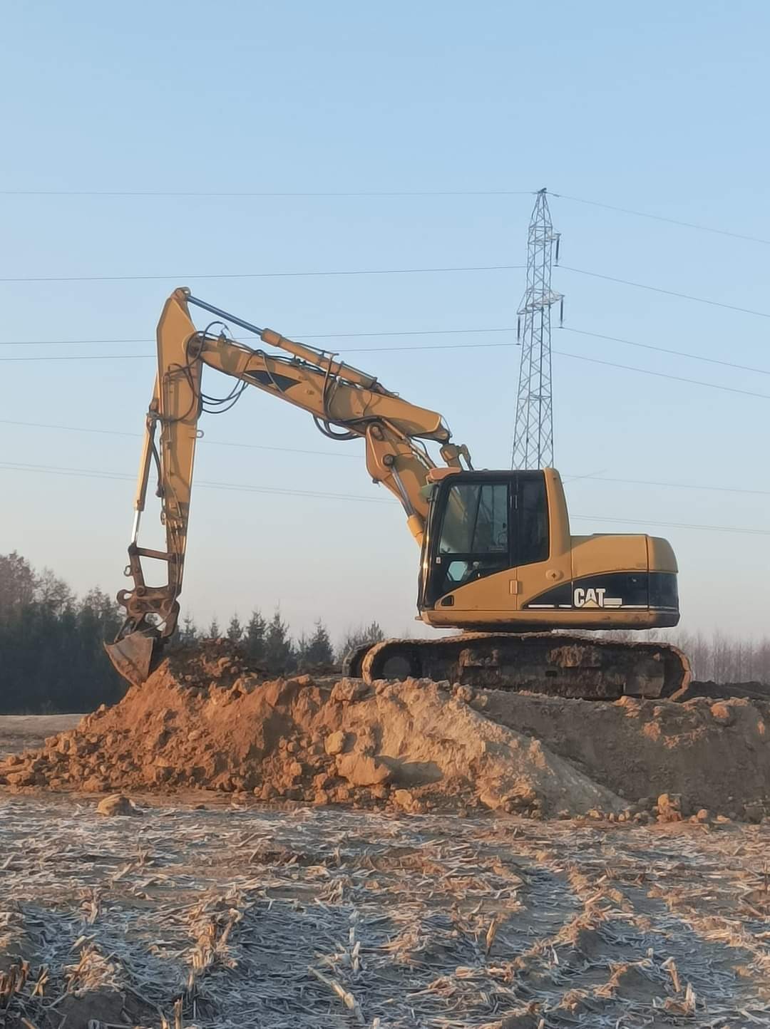Żółta koparka CAT nabiera ziemię na polu z resztkami po żniwach, w tle linia wysokiego napięcia i drzewa.