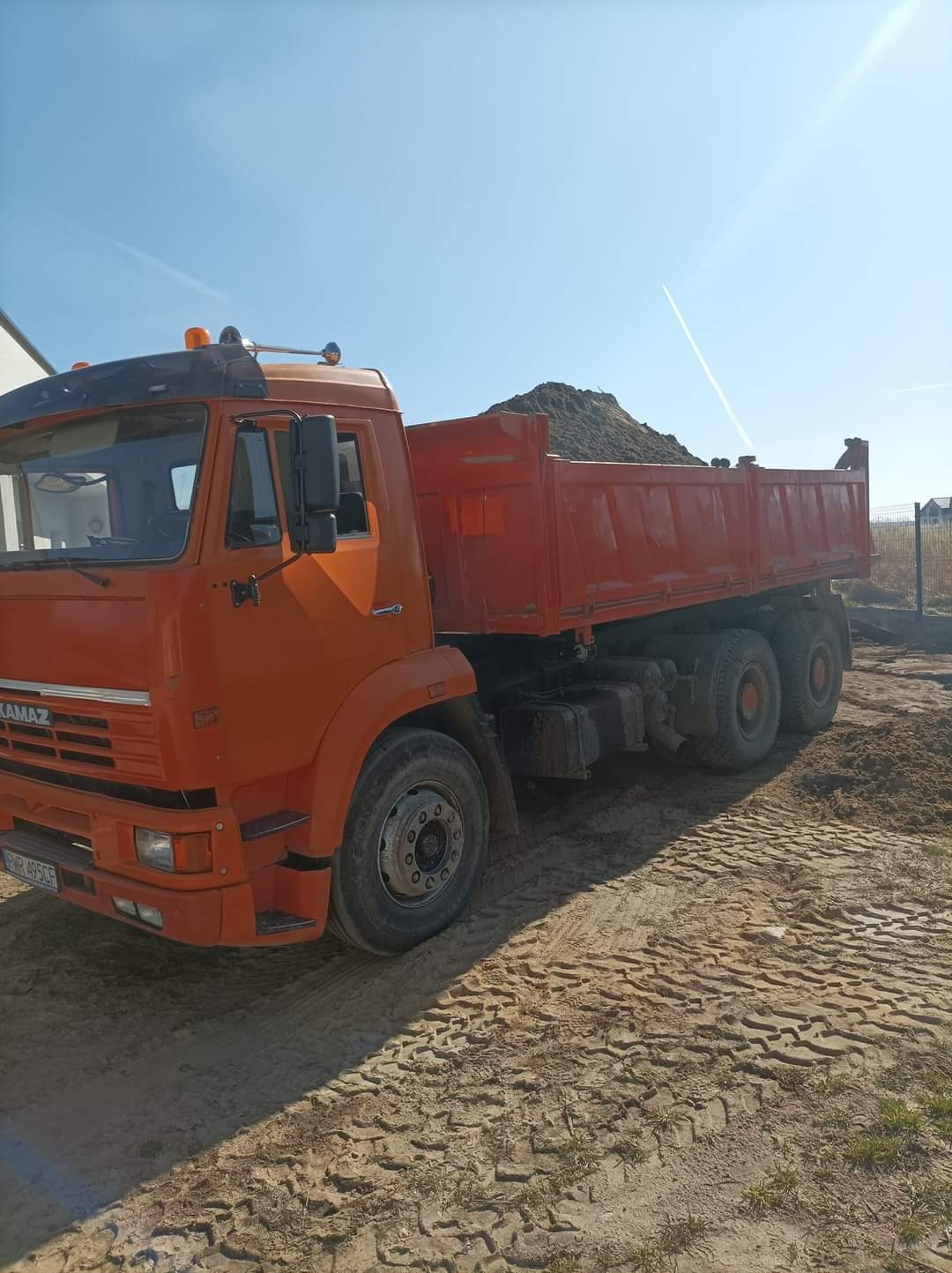 Pomarańczowa ciężarówka wywrotka marki Kamaz z ładunkiem ziemi na pace, stojąca na piaszczystym terenie w słoneczny dzień.