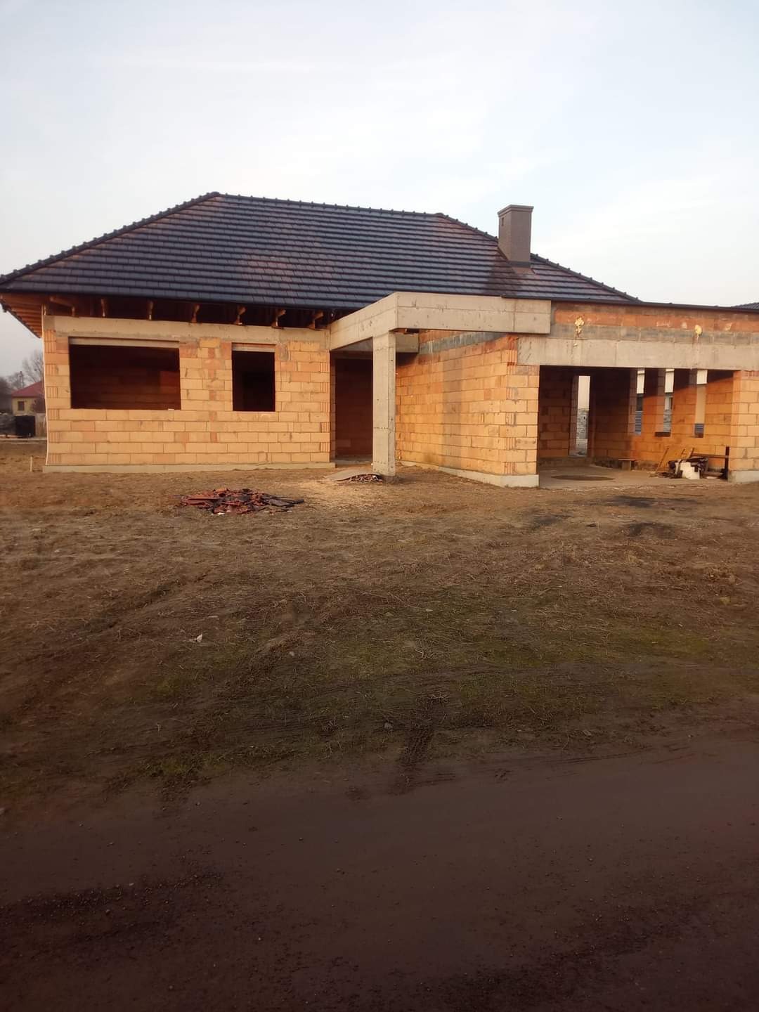 Niedokończony dom jednorodzinny z ciemną dachówką, widoczne otwory okienne i drzwiowe, ściany z pustaków ceramicznych, teren wokół niezagospodarowany, w tle zabudowa jednorodzinna.