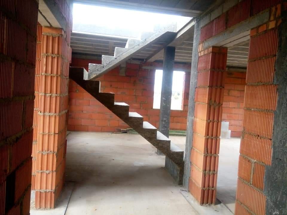 Wnętrze domu w trakcie budowy z widocznymi ścianami z pustaków ceramicznych, betonowymi schodami wewnętrznymi oraz oknami bez ram.