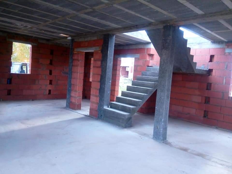Wnętrze budynku w trakcie budowy: ściany z czerwonej cegły dziurawki, betonowe schody na piętro wsparte na słupie, surowy betonowy strop, widoczne otwory okienne i drzwiowe.