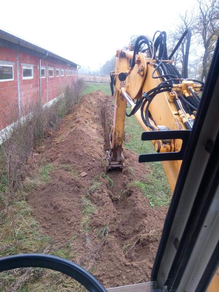 Żółta koparka z kabiny, wykonująca wąski wykop w ziemi wzdłuż ściany budynku z czerwonej cegły, widoczne przewody hydrauliczne i osprzęt roboczy.