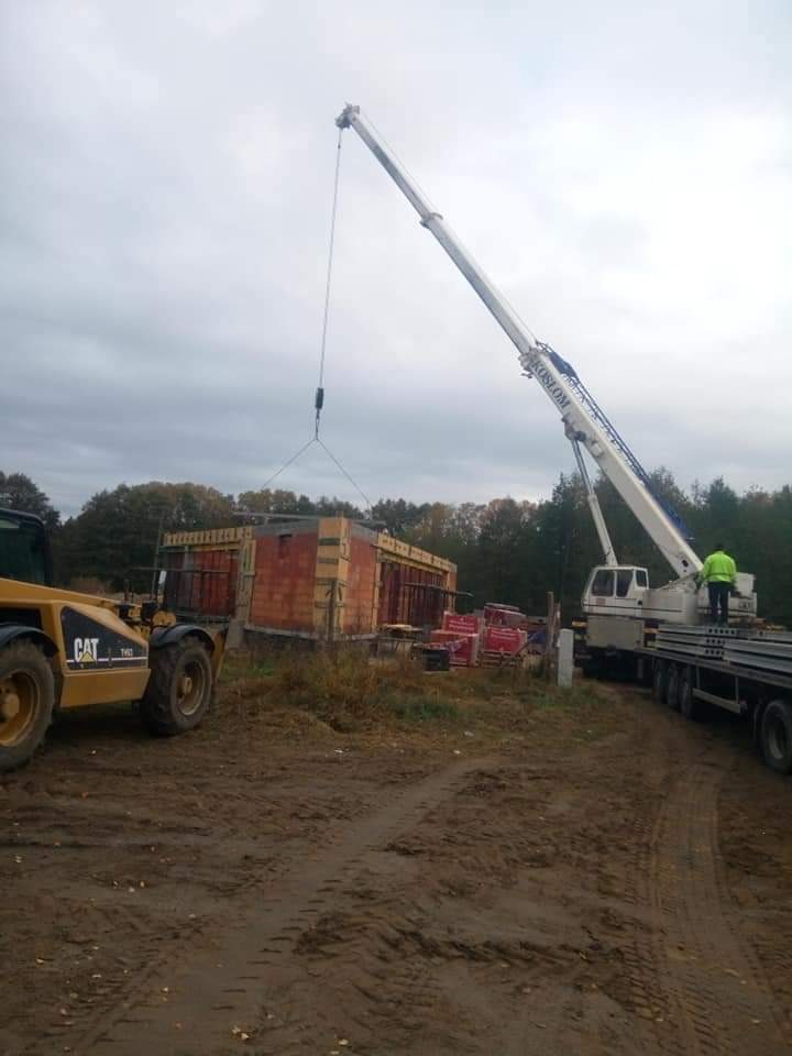 Budowa domu: żuraw przenosi element konstrukcyjny na placu budowy, widoczny fragment ściany z czerwonej cegły i żółta ładowarka CAT na błotnistym terenie.