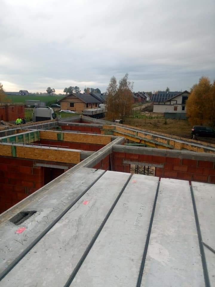 Budowa domu w trakcie: widok z góry na wznoszone ściany z czerwonej cegły i ułożone płyty stropowe z betonu z widocznymi napisami, drewniane szalunki w tle, krajobraz wiejski w oddali...