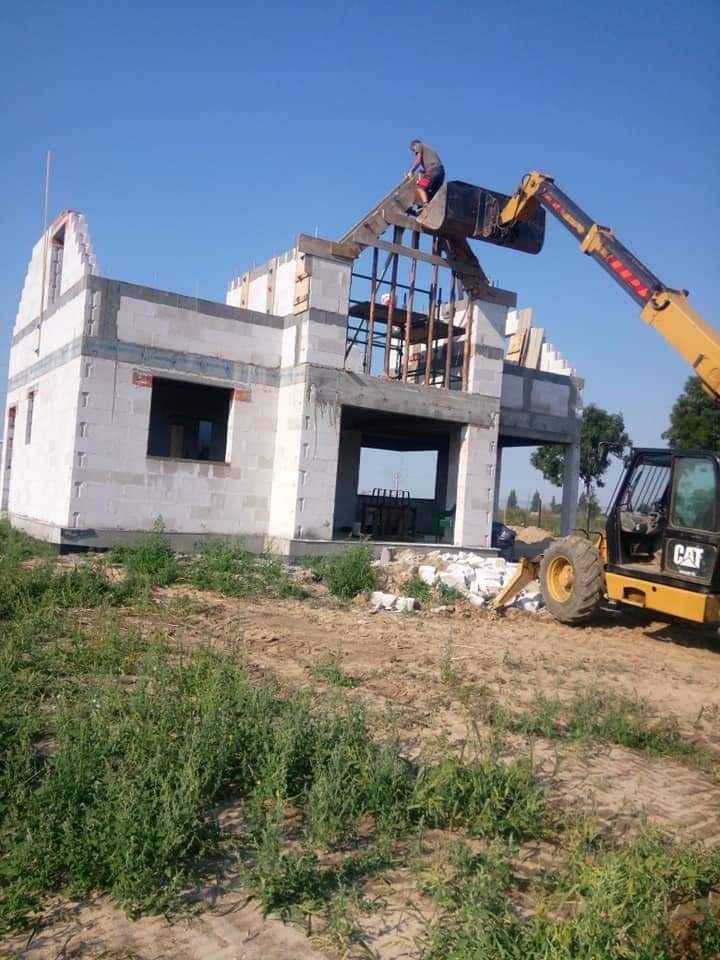 Wznoszenie dwukondygnacyjnego domu z białego bloczka z użyciem ładowarki teleskopowej CAT, pracownik na dachu układa elementy konstrukcyjne, w tle błękitne niebo.