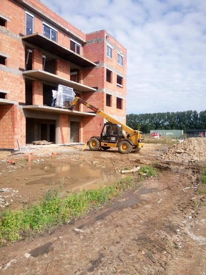 Żółta ładowarka teleskopowa podnosi okna na balkon w surowym stanie budynku z czerwonej cegły, teren budowy jest błotnisty po deszczu.