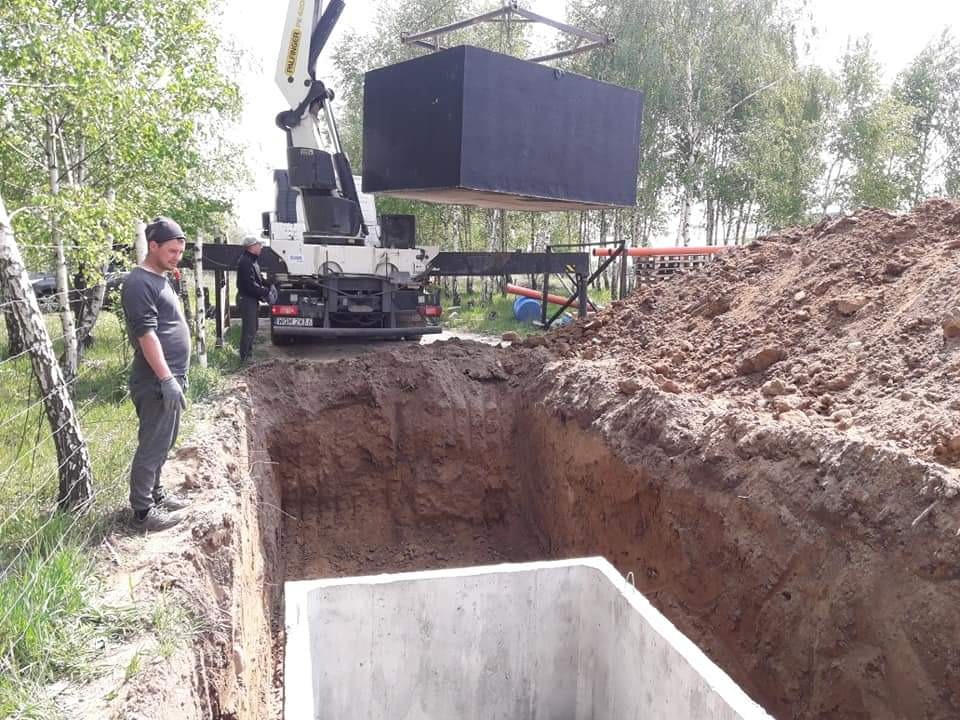 Montaż betonowego zbiornika w wykopie za pomocą dźwigu Palfinger, dwóch pracowników nadzoruje proces, widoczny stos ziemi i fragment ogrodzenia z siatki.