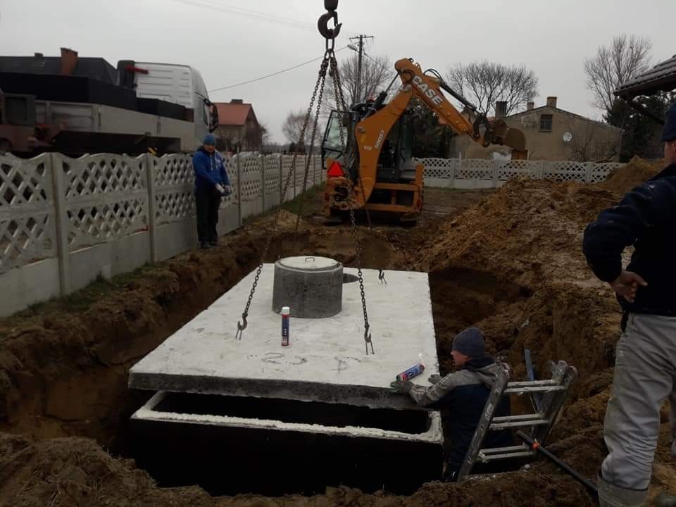 Montaż betonowego zbiornika pod ziemią przy użyciu koparki i dźwigu; pracownik na drabinie uszczelnia połączenie elementów zbiornika.