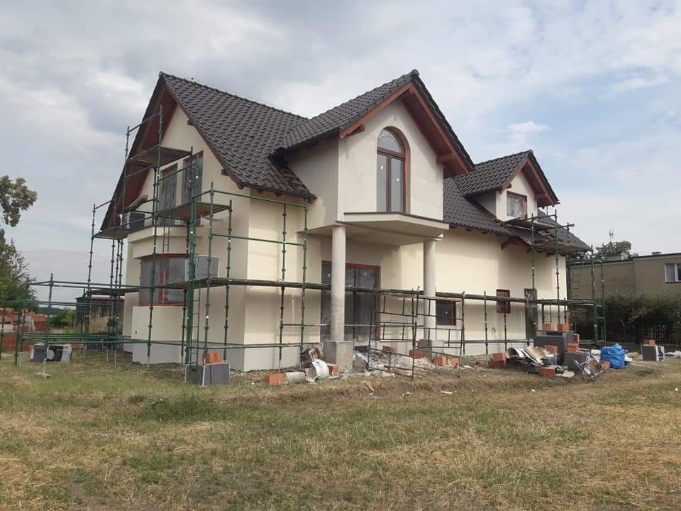 Niedokończony dom z jasną elewacją w trakcie budowy, otoczony rusztowaniami, z widocznymi cegłami i materiałami budowlanymi na trawniku.
