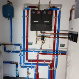 Nowoczesna kotłownia z piecem CO, zbiornikiem wyrównawczym i systemem rur w kolorach niebieskim i czerwonym, zamocowanych na białej ścianie.