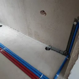 Surowy stan: Instalacja wodna z rurami w kolorze niebieskim i czerwonym, częściowo zamontowana w surowej ścianie i podłodze, widoczne otwory w ścianie i podłodze, szare rury kanalizacyjne.
