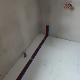 Instalacja podłogowa z rurami grzewczymi w bruzdach w styropianie, widoczne wystające rury ze ściany i puszki elektryczne.