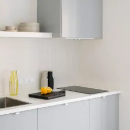 Jasna, minimalistyczna kuchnia z szarymi szafkami, białym blatem i płytkami, z czarną deską do krojenia z połówkami cytryny, butelką z żółtymi paskami i czarną płytą indukcyjną.