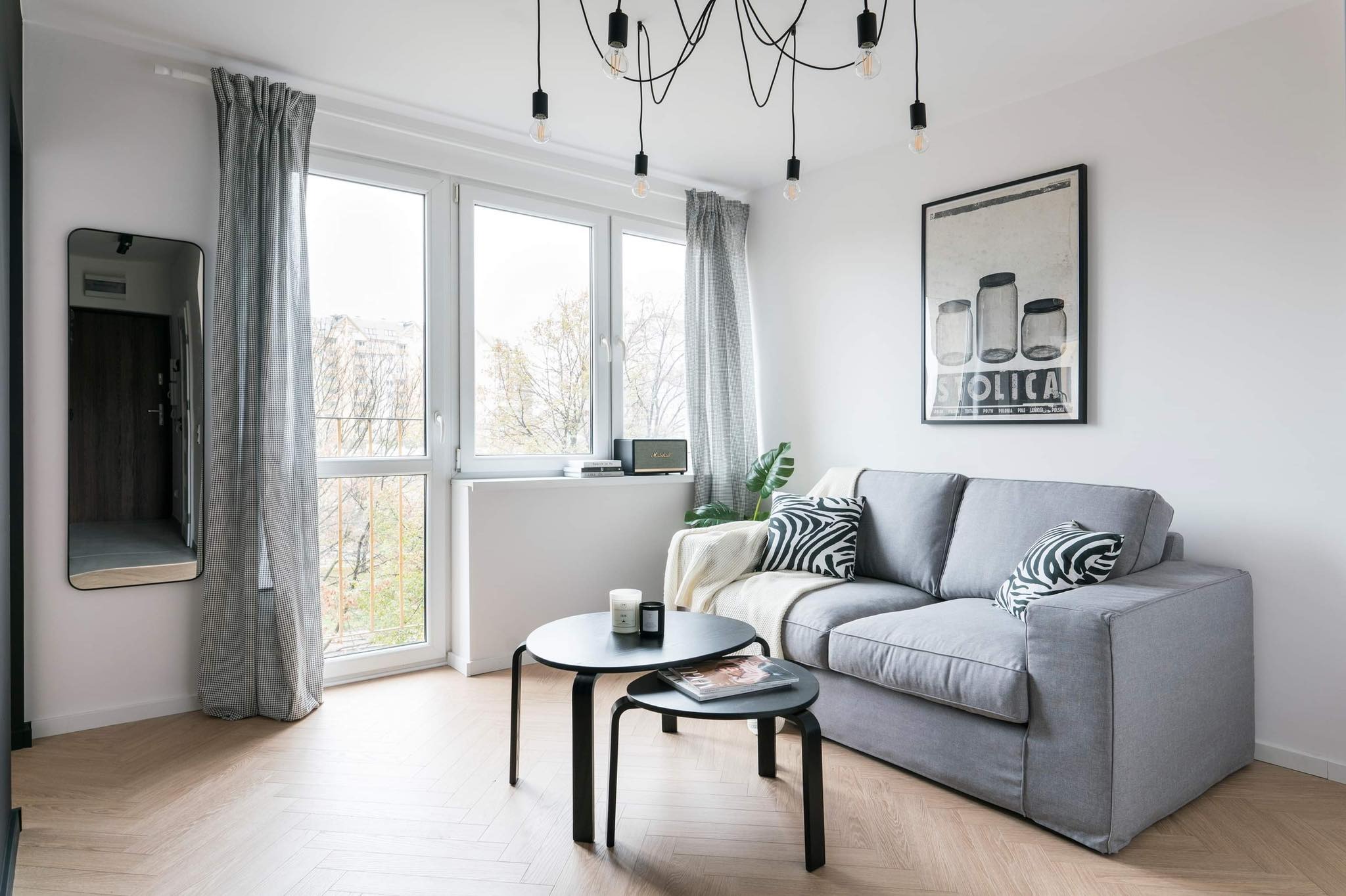 Jasne wnętrze pokoju dziennego z szarą sofą, stolikami kawowymi, oknem z widokiem na drzewa i lampą wiszącą z odsłoniętymi żarówkami.