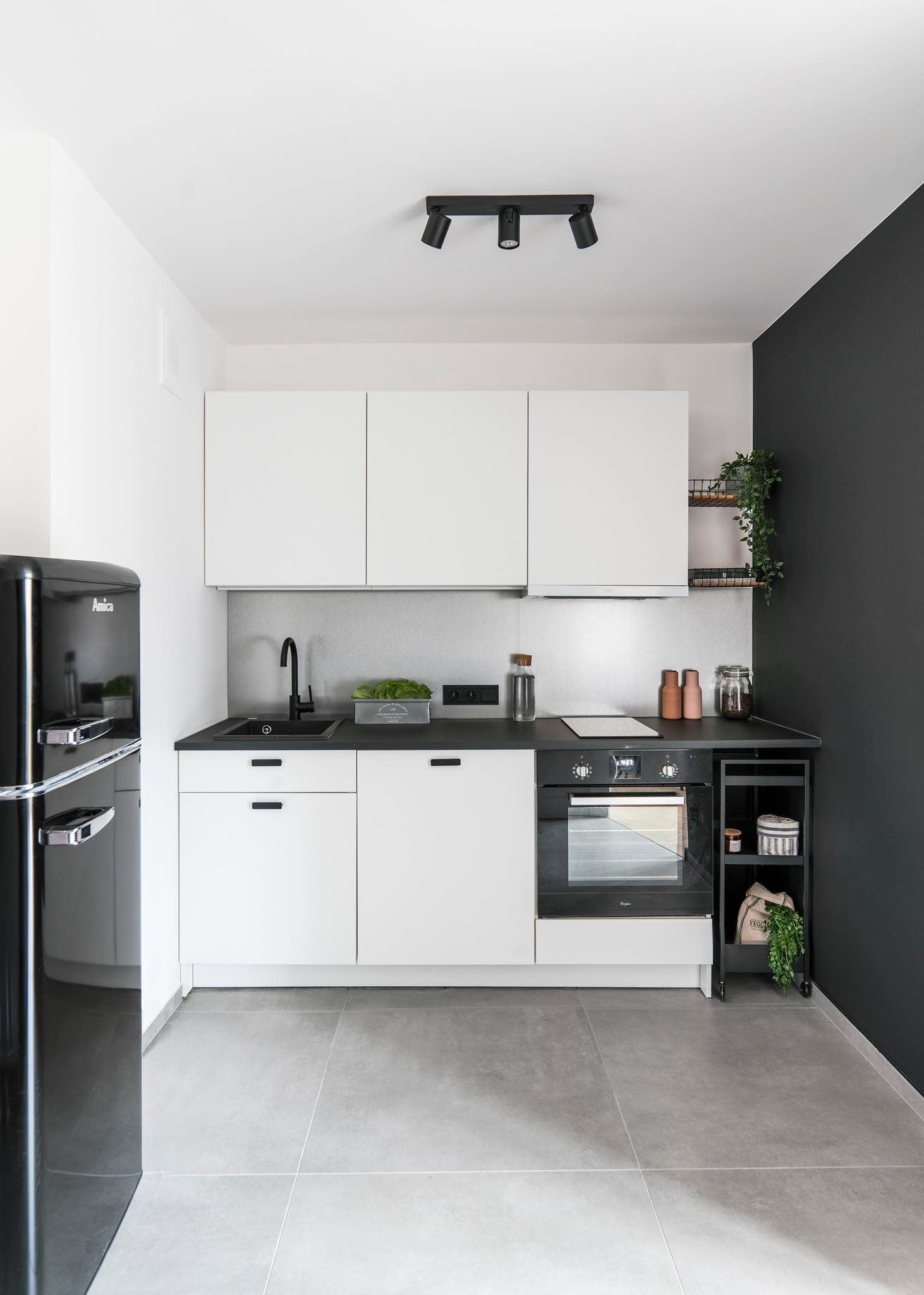 Nowoczesna, minimalistyczna kuchnia z czarnym retro lodówką Amica, białymi szafkami, czarnym blatem i ścianą pomalowaną na ciemny grafit. Na blacie zioła w metalowej doniczce i szklane pojemniki.