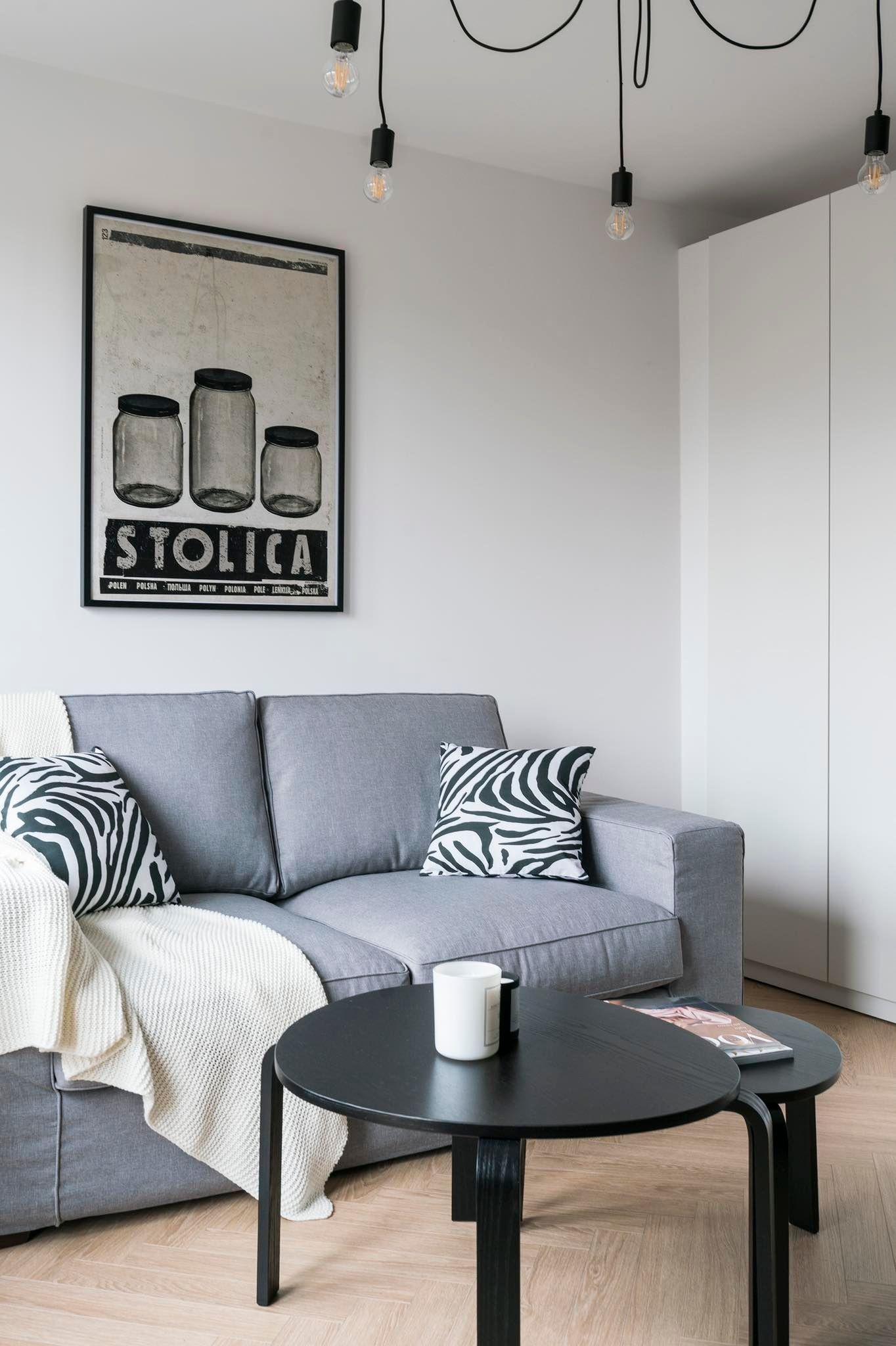 Szary narożnik z poduszkami w zebrę i kremowym pledem, czarne okrągłe stoliki kawowe, plakat z motywem słoików nad sofą, minimalistyczne oświetlenie z żarówkami Edisona, parkiet w jodełkę.