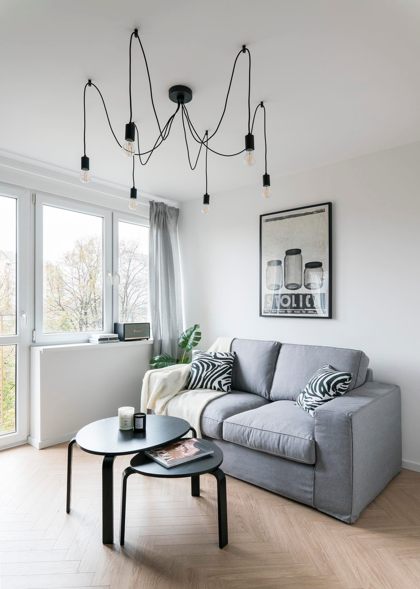 Jasne wnętrze pokoju dziennego z szarą sofą, czarnymi stolikami kawowymi, lampą pająk i parkietem ułożonym w jodełkę, oświetlone naturalnym światłem z okna.