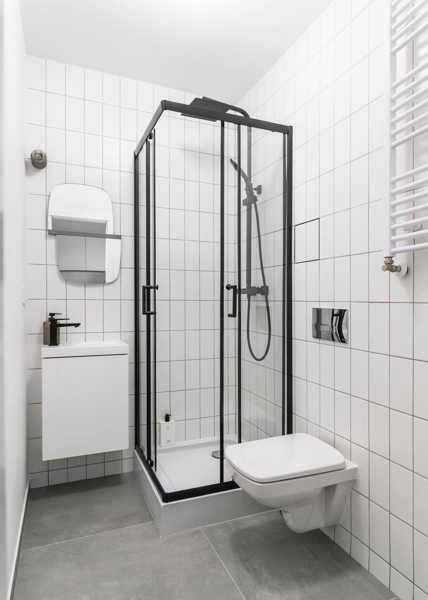 Minimalistyczna łazienka z białymi kwadratowymi płytkami, czarną kabiną prysznicową, podwieszaną toaletą i umywalką z czarną armaturą, uzupełniona grzejnikiem drabinkowym i lustrem z półką.