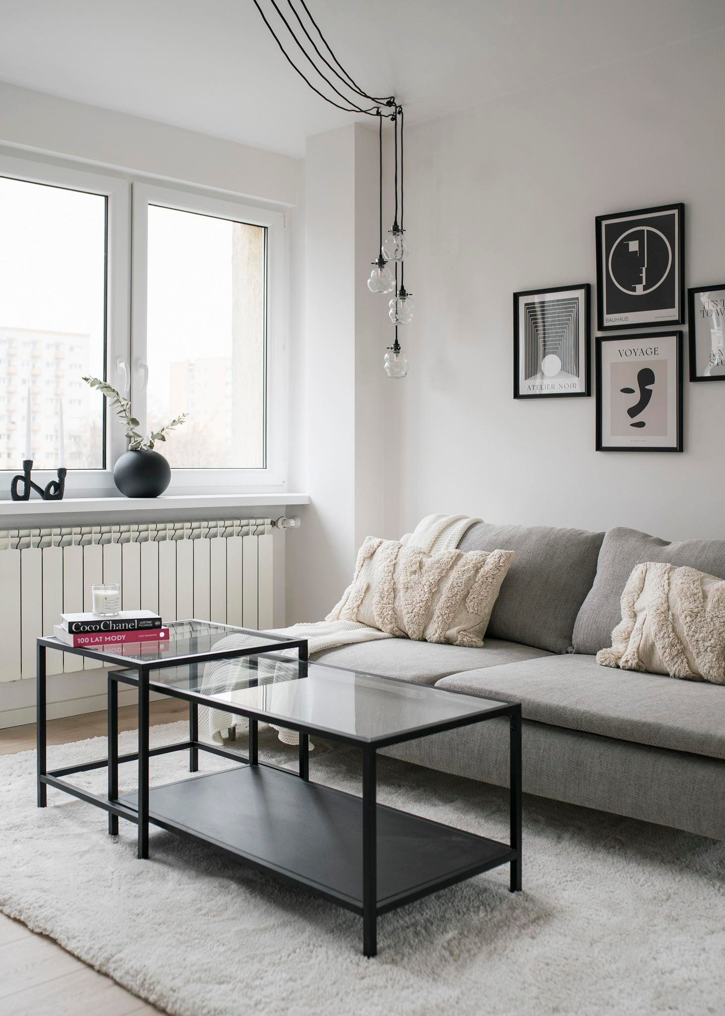Jasne wnętrze salonu z szarą sofą, czarnym stolikiem kawowym z szklanym blatem, dekoracyjnymi poduszkami i minimalistycznymi grafikami na ścianie.