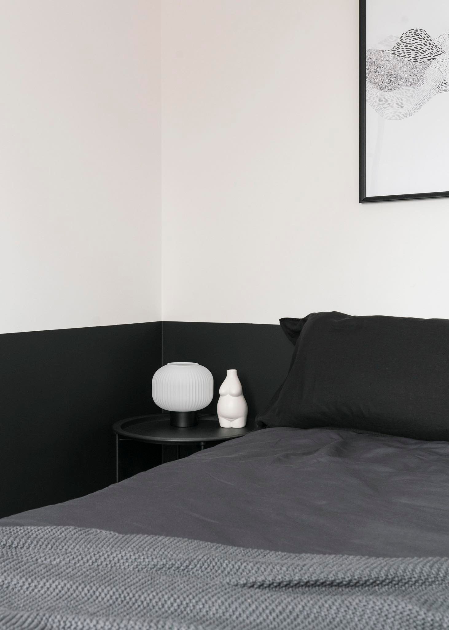 Minimalistyczna sypialnia w Warszawie z czarno-białymi ścianami, szarą pościelą i lampką nocną na czarnym stoliku, uzupełniona abstrakcyjnym obrazem w czarnej ramie.