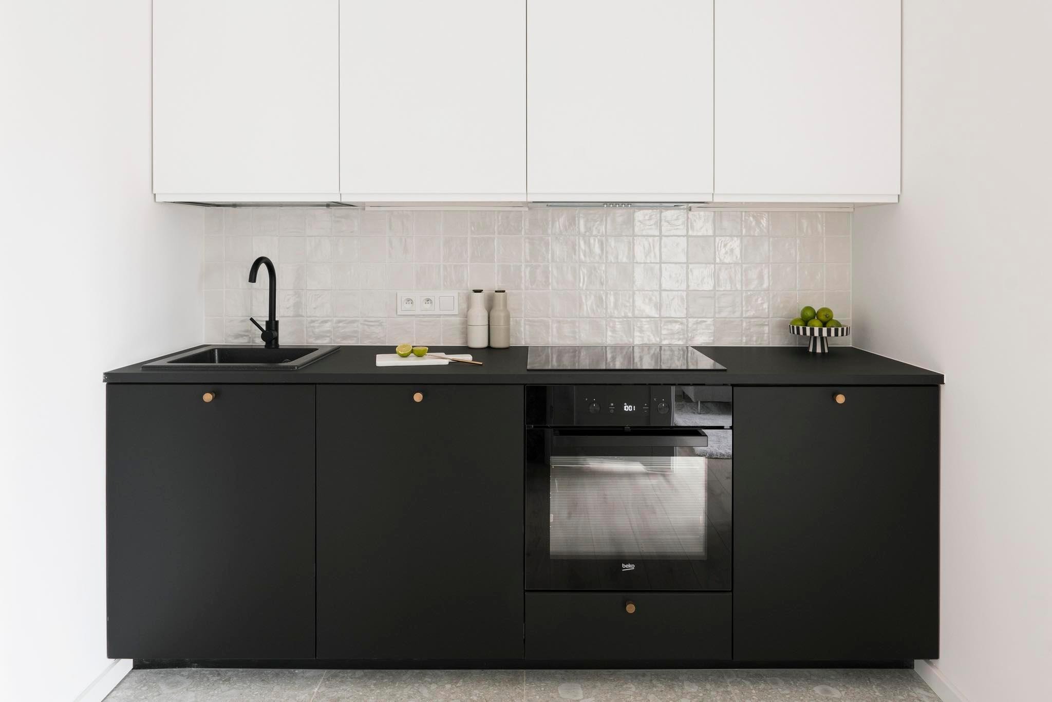 Minimalistyczna kuchnia z czarnymi szafkami i białymi górnymi szafkami, płytki ceramiczne z delikatnym wzorem, czarna armatura i drewniane gałki, limonki na desce do krojenia.