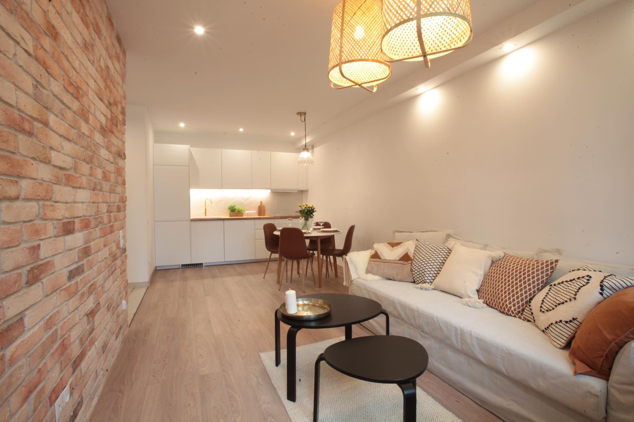 Jasne wnętrze mieszkania z ceglaną ścianą, drewnianą podłogą, białymi szafkami kuchennymi, okrągłym stołem z krzesłami, sofą z dekoracyjnymi poduszkami i dwoma okrągłymi stolikami kawowymi na dywanie.