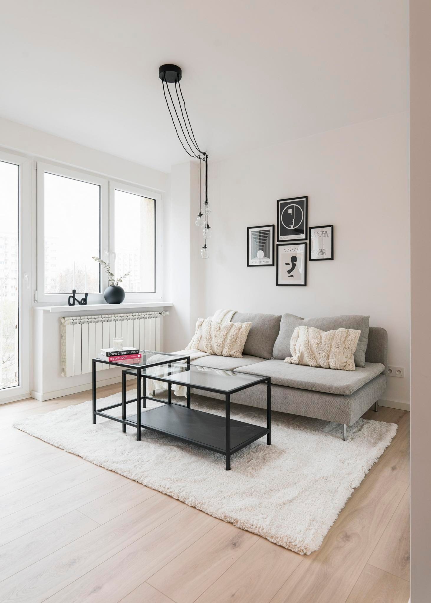 Jasne wnętrze salonu z szarą sofą, puszystym dywanem, czarnym stolikiem kawowym z szklanym blatem i galerią czarno-białych grafik na ścianie, wisząca lampa z trzema żarówkami.