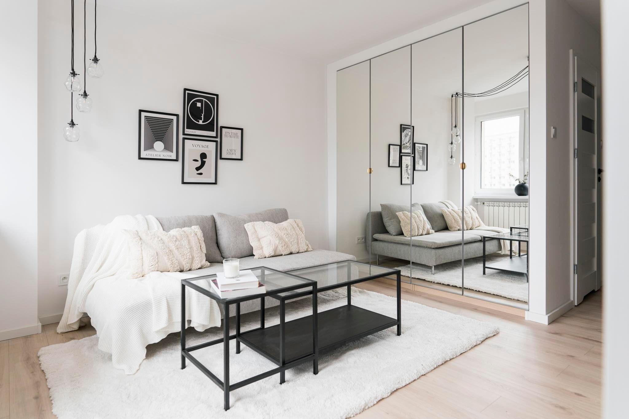 Jasne wnętrze salonu z szarą sofą, białym dywanem shaggy, stolikiem kawowym z czarną ramą i szklanym blatem, oraz lustrzaną szafą w tle, odbijającą okno i część pokoju.