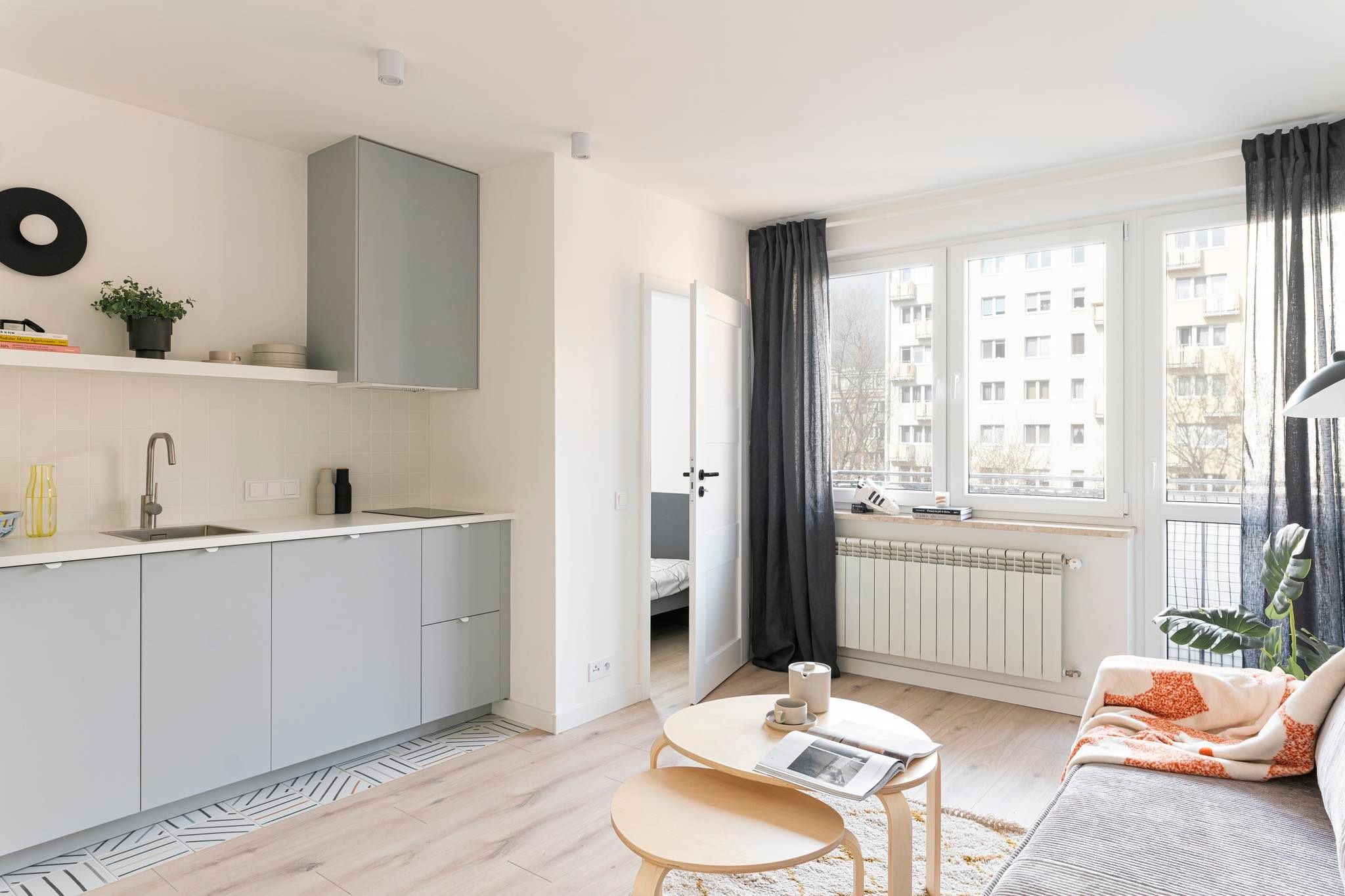 Jasne wnętrze mieszkania z aneksem kuchennym i widokiem na sypialnię, szare szafki kuchenne, drewniany stół kawowy i sofa z wzorzystym pledem, okno z szarymi zasłonami.