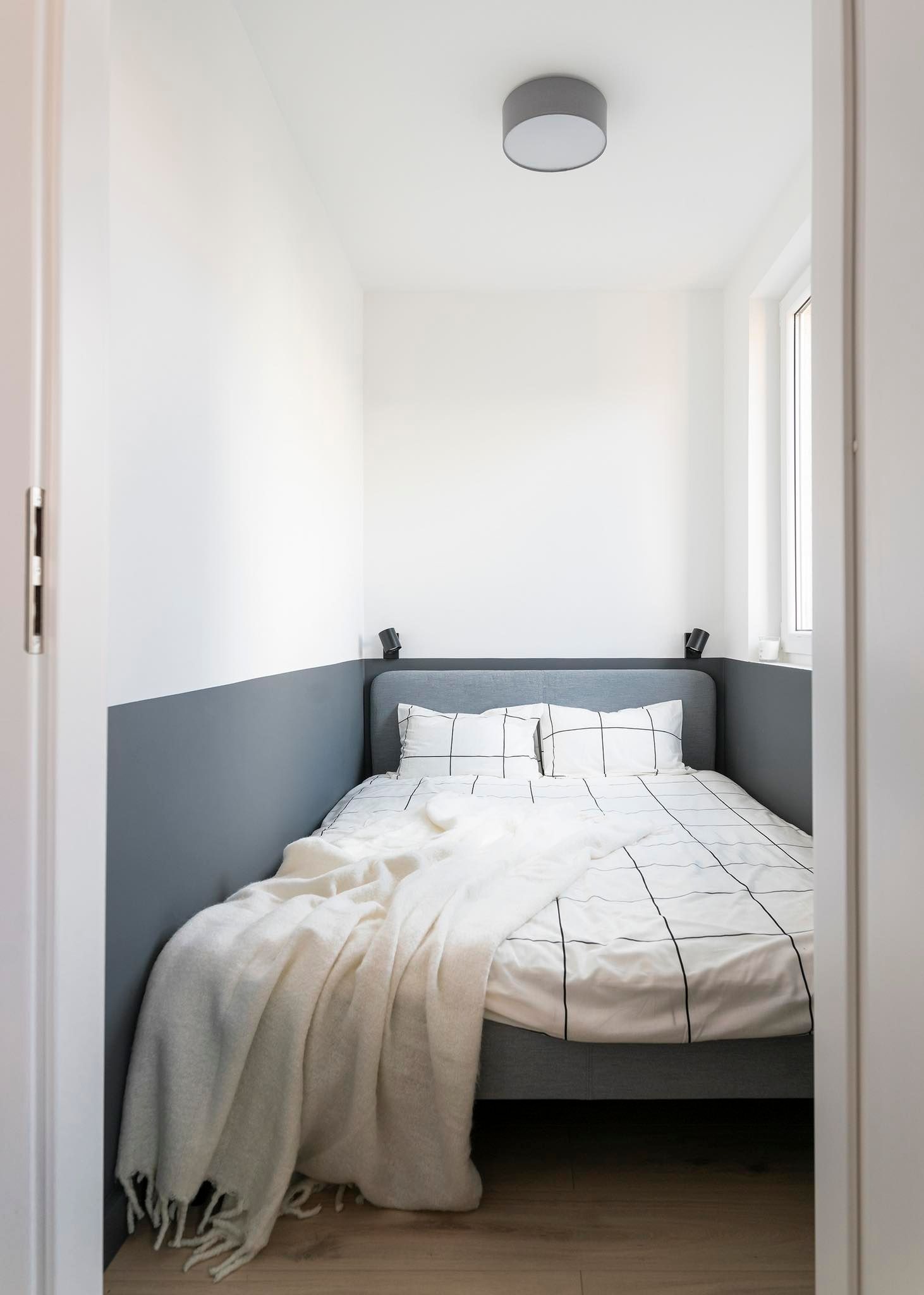 Minimalistyczna sypialnia z łóżkiem w szarej ramie, białą pościelą w czarną kratę i kremowym pledem, ściany pomalowane na biało i szaro, z drewnianą podłogą i oświetleniem punktowym.