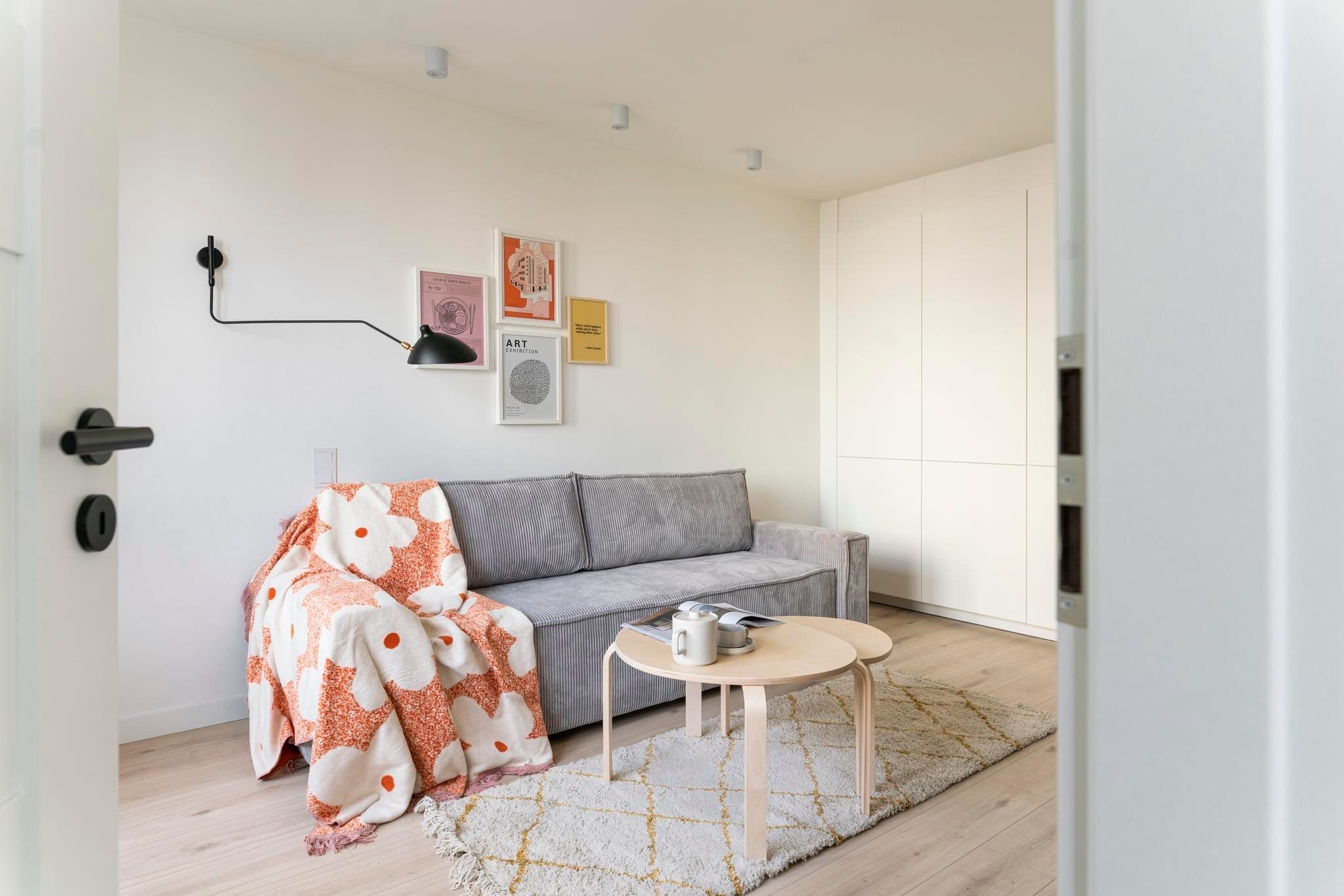 Jasne wnętrze pokoju dziennego z szarą sofą, okrągłymi stolikami kawowymi i wzorzystym pledem w kwiaty, oświetlone czarną lampą ścienną i punktowymi reflektorami sufitowymi.
