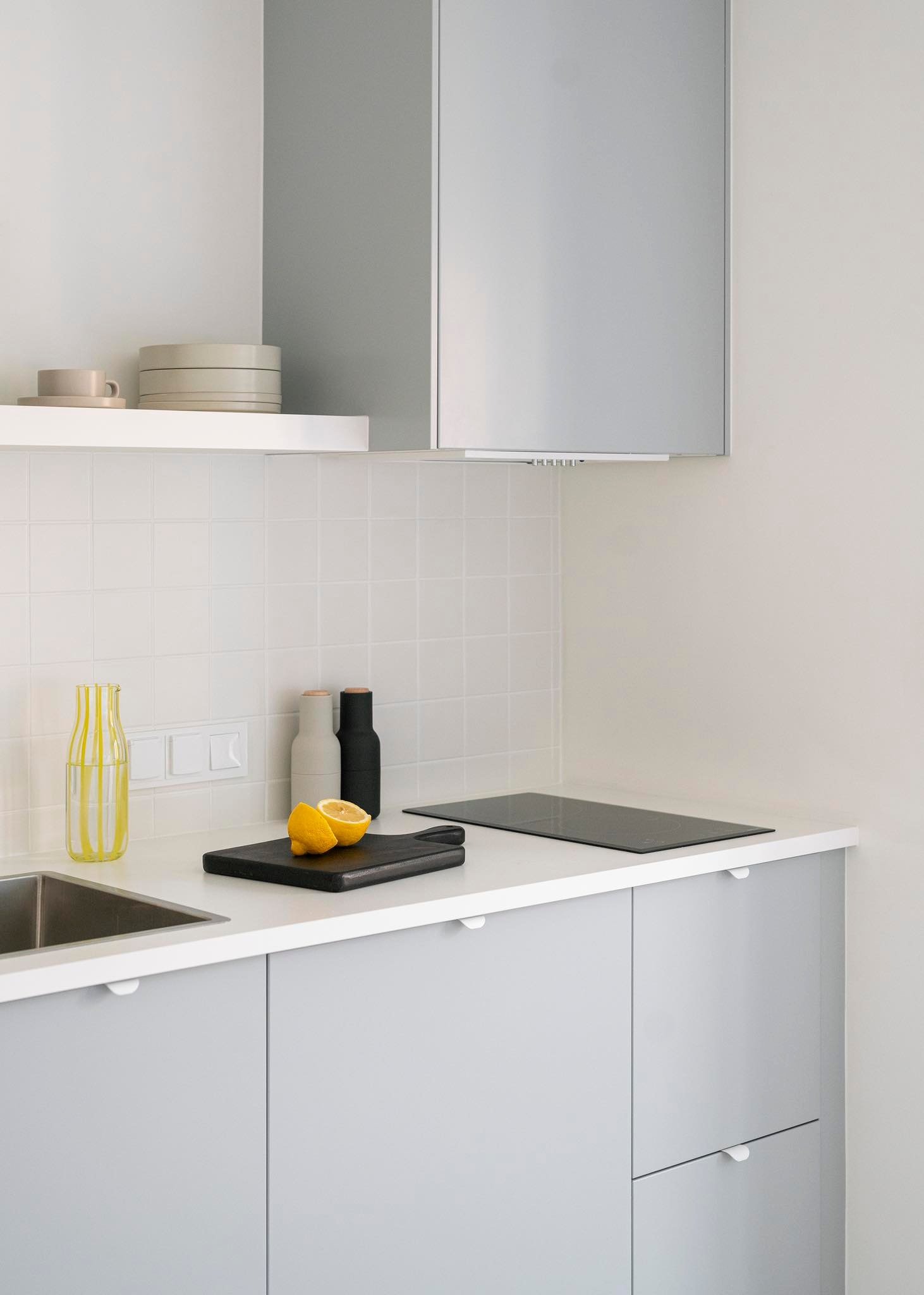 Jasna, minimalistyczna kuchnia z szarymi szafkami, białym blatem i płytkami, z czarną deską do krojenia z połówkami cytryny, butelką z żółtymi paskami i czarną płytą indukcyjną.