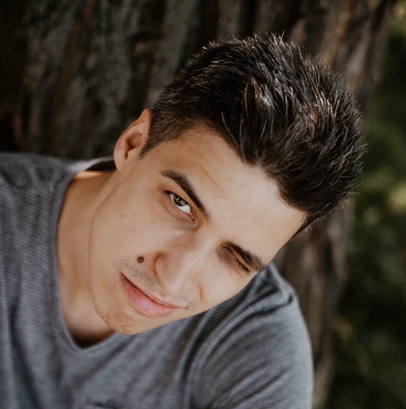 Portret młodego mężczyzny z przymrużonym okiem, opierającego się o korę drzewa, z widocznym pieprzykiem nad ustami.