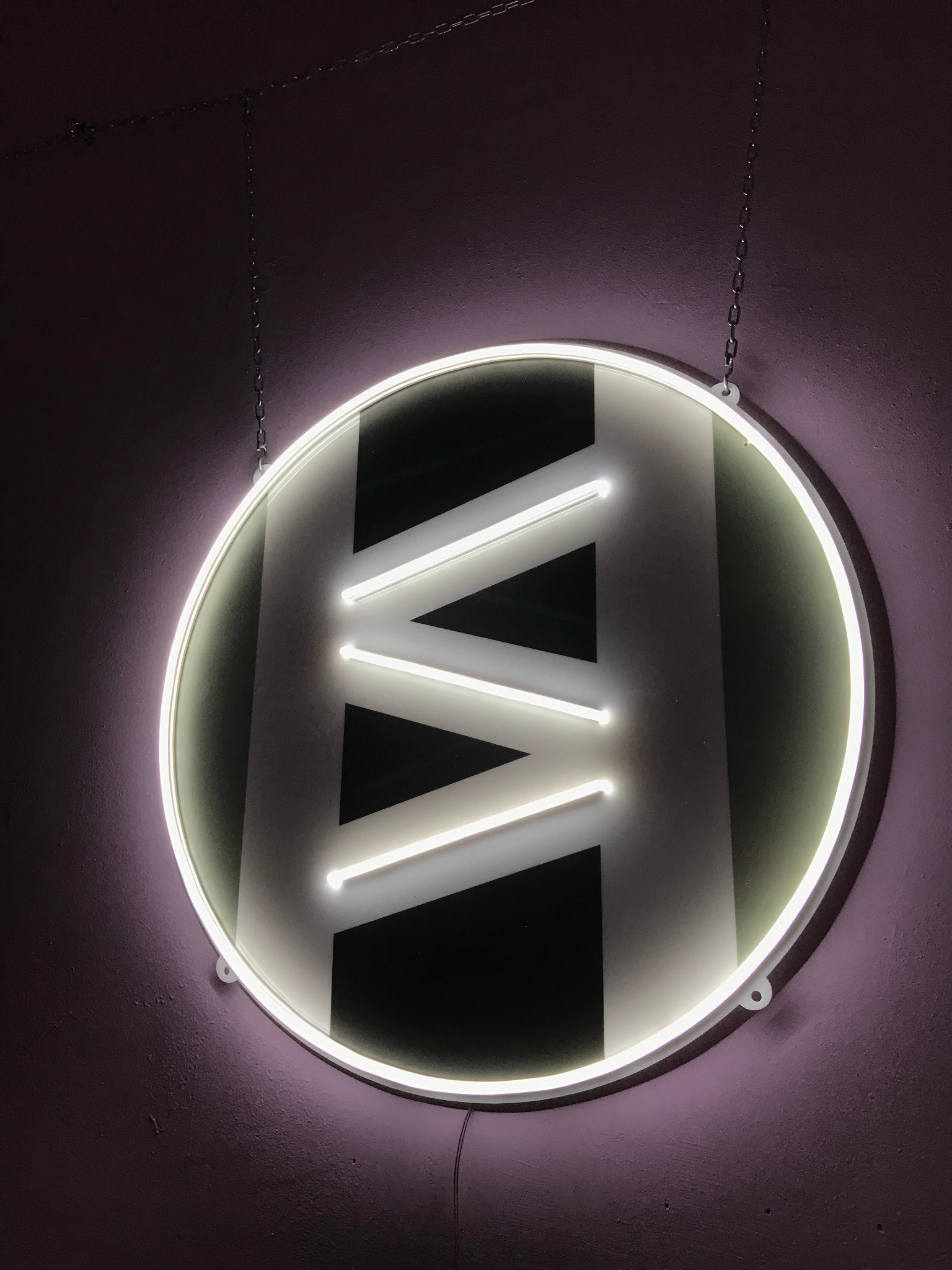 Podświetlane neonowe logo w kształcie litery H z trzema poziomymi kreskami wewnątrz, zawieszone na łańcuszku na fioletowej ścianie.