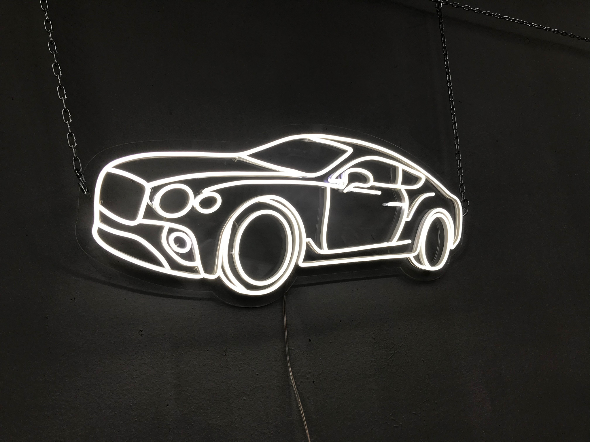 Neonowy kontur sportowego samochodu marki Bentley zawieszony na łańcuchach na ciemnej ścianie, z widocznym kablem zasilającym.
