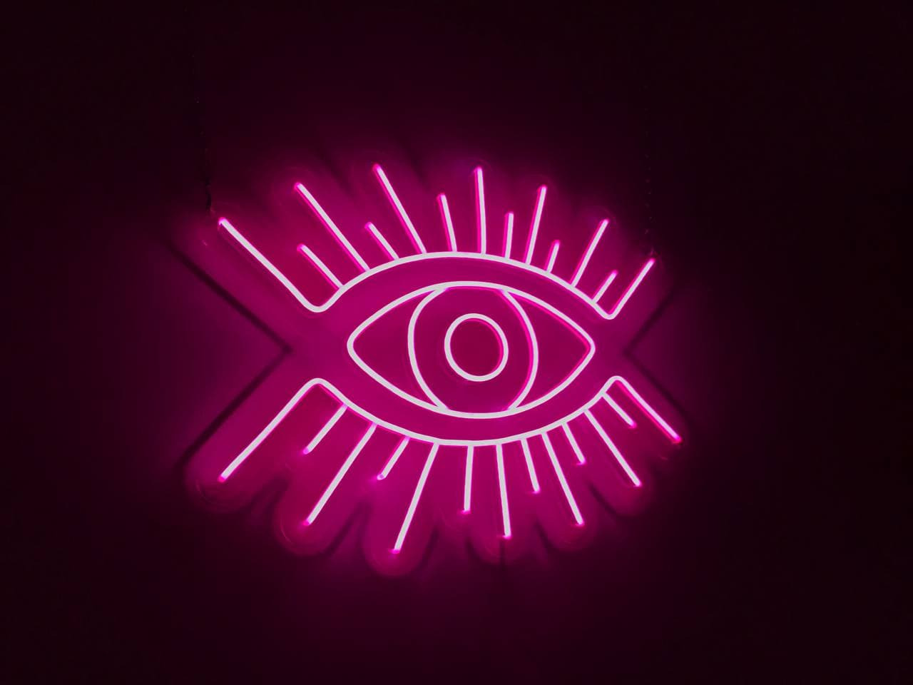 Neonowy znak w kształcie oka z promienistymi liniami, świecący intensywnym różowym światłem na ciemnym tle.