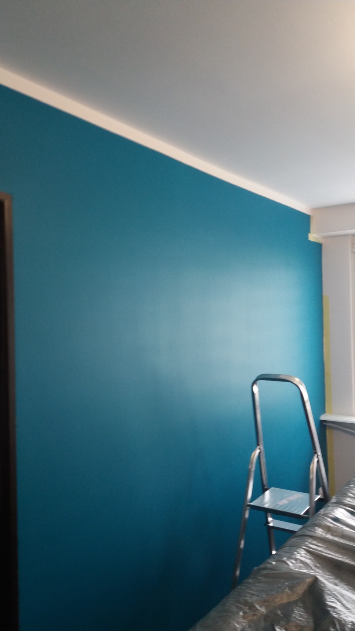 Świeżo pomalowana na turkusowo ściana w pokoju, widoczna aluminiowa drabina i folia ochronna na podłodze, biały sufit z listwą.