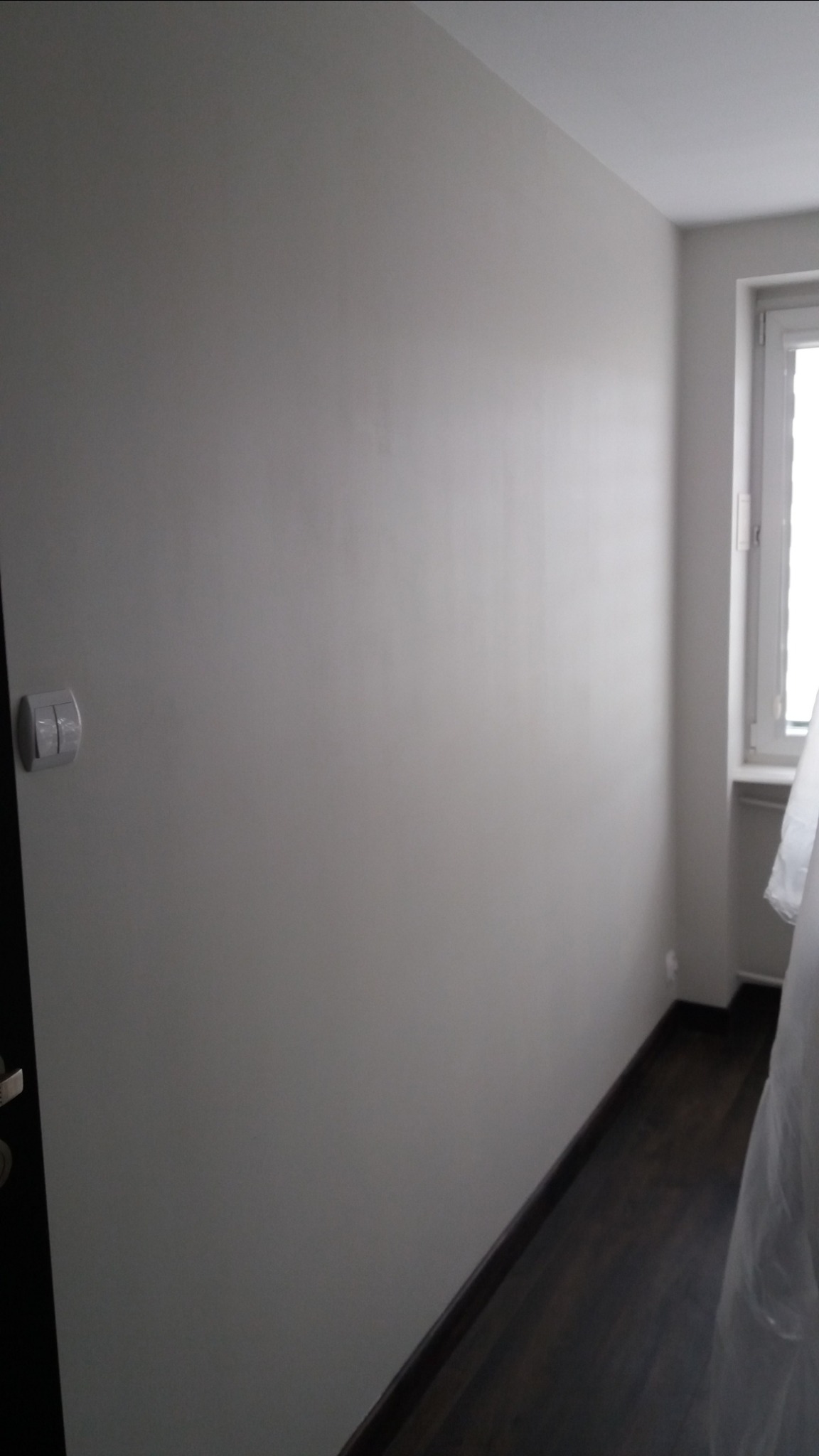 Świeżo pomalowana ściana w odcieniach szarości z widocznym włącznikiem światła, ciemna podłoga i fragment okna z zasłoniętą szybą.