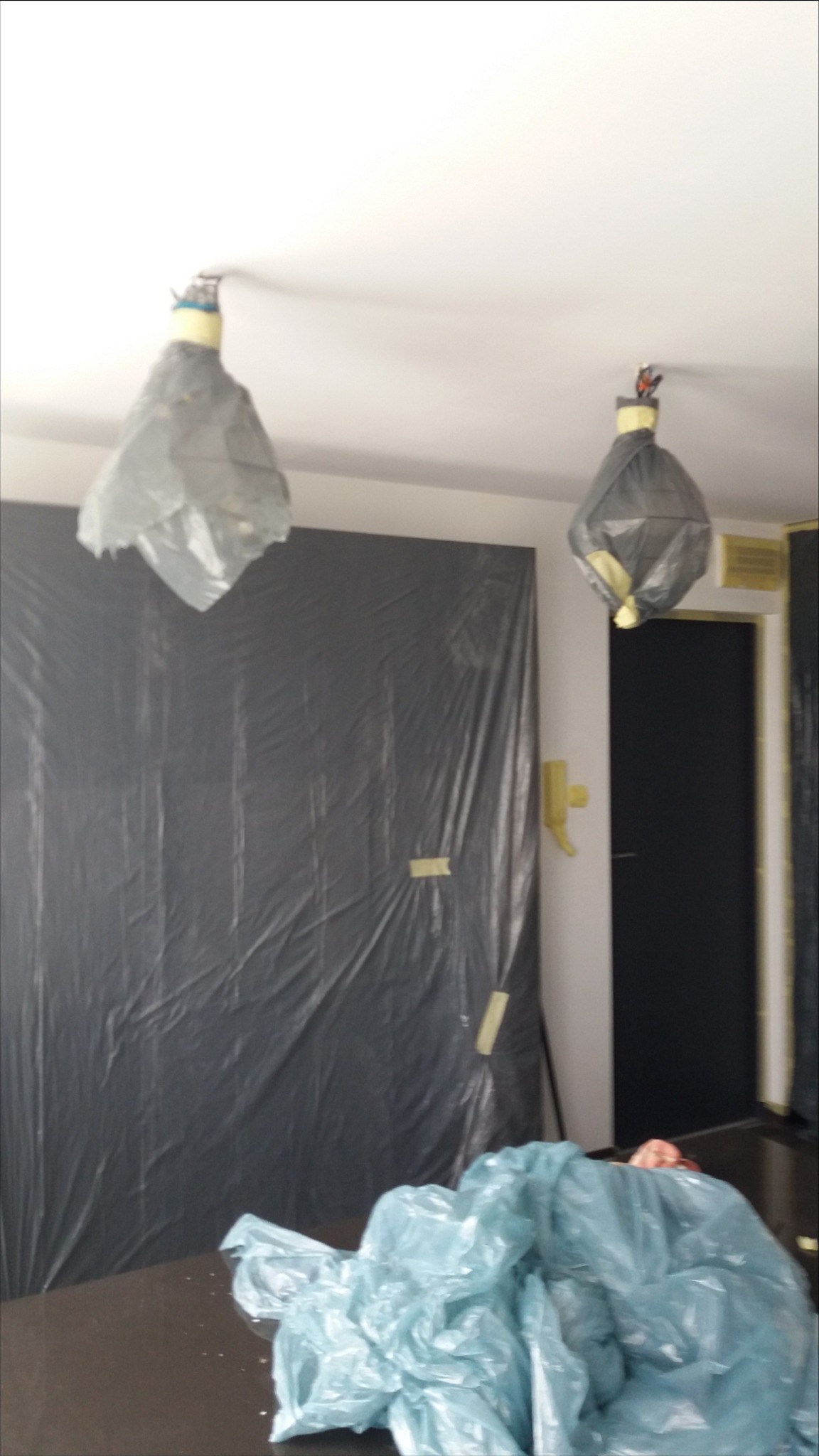 Przygotowanie do malowania: zabezpieczone lampy folią i ściana zakryta czarną folią malarską, na stole niebieska folia ochronna.