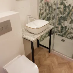 Nowoczesna łazienka z umywalką nablatową na blacie imitującym kamień, kabiną prysznicową z motywem roślinnym i toaletą podwieszaną, wykończona płytkami w odcieniach bieli i drewna.